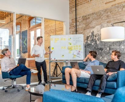 Agile three men sitting while using laptops and watching man beside whiteboard